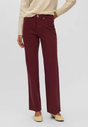 Femme portant un pantalon droit taille haute rouge foncé, un pull en maille beige et des chaussures beiges à petits talons, debout avec une main sur la hanche.