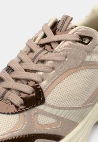 Chaussure de sport beige avec une tige en mesh, des accents marron brillants, des lacets texturés et une semelle légère avec un motif subtil.