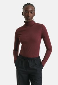 Frau mit geflochtenem Haar, die einen figurbetonten burgunderfarbenen Rollkragenpullover und schwarze elastische Hosen trägt, steht mit den Händen in den Taschen vor einem weißen Hintergrund.