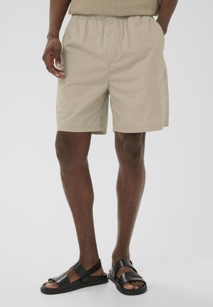Mand iført beige knælange shorts og sorte lædersandaler, stående med den ene hånd i lommen mod en ensfarvet baggrund.
