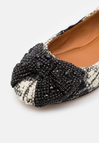 Chaussure plate avec nœud noir perlé à l'avant et dessus en tissu tissé noir et blanc texturé, doublure intérieure couleur fauve visible.