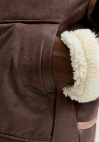 Ręka osoby w kieszeni brązowej kurtki z owczej wełny z widoczną miękką, kremową podszewką na mankietach.