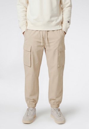 Hombre vestido con pantalones cargo beige con puños elásticos, bolsillos laterales, zapatillas blancas y sudadera color crema con las manos en los bolsillos.