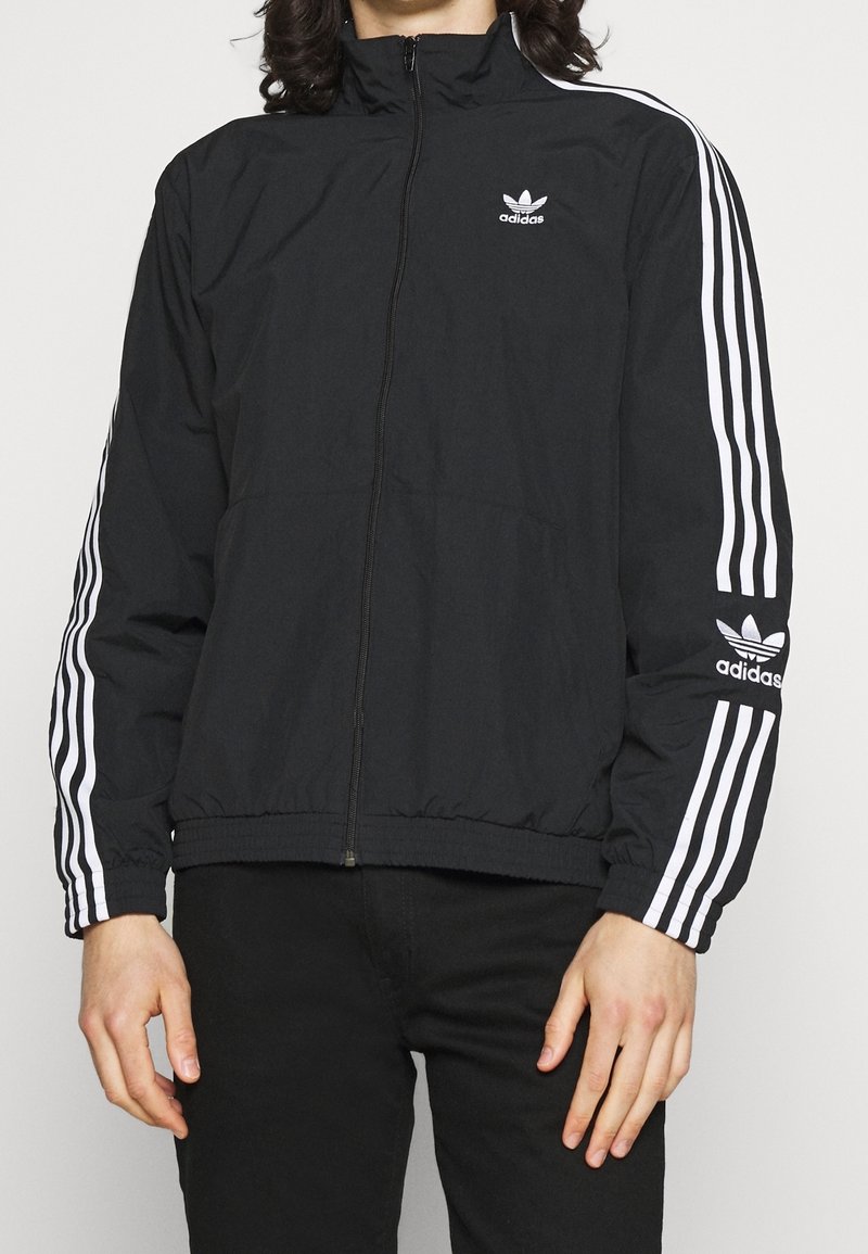 Musta adidas-zip-up-takki, jossa valkoiset kolme raitaa korostavat hihoja ja logo rintakehällä. Kevyt, rento malli, jossa joustavat hihansuut ja helma.