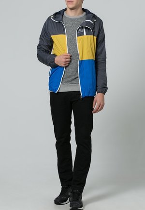 Veste color-block en bleu, jaune et gris, avec une texture légère et lisse. Dotée d'une capuche et d'une fermeture éclair, assortie à un pantalon noir.