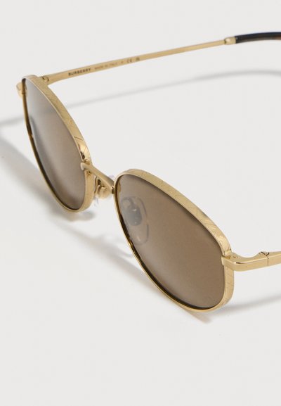 Στρογγυλά γυαλιά ηλίου με χρυστό μεταλλικό πλαίσιο, καφέ φακούς με απόχρωση και ρυθμιζόμενα μαξιλαράκια μύτης, τοποθετημένα σε λευκή επιφάνεια.