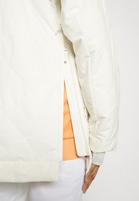 Gestepptes, cremeweißes Jackett mit seitlichen Reißverschlüssen, das ein strukturiertes Design zeigt. Ein orangefarbener Sweatshirt ist darunter mit einem gerippten Saum sichtbar.