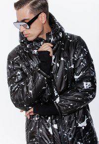 Młody mężczyzna poprawiający kaptur czarnej kurtki puchowej w biały wzór plam farby, noszący czarne okulary przeciwsłoneczne, na białym tle.