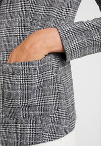 Zwart-wit houndstooth patroon stof met een zijzakdetail. Gladde textuur, op maat gemaakte ontwerp, en lange mouwen zichtbaar.