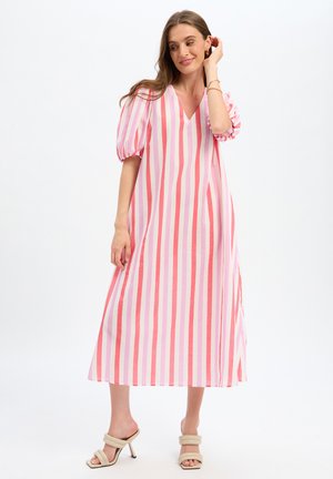 Donna che indossa un abito midi a righe verticali rosa e bianche con maniche a sbuffo e sandali beige con tacco, posa con una mano vicino al viso.