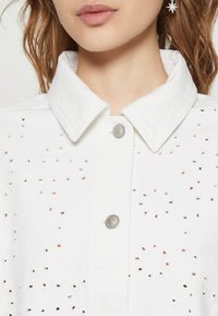 Chaqueta de mezclilla blanca con intrincados patrones recortados, un cuello puntiagudo y botones a presión plateados. Textura suave con acentos florales.