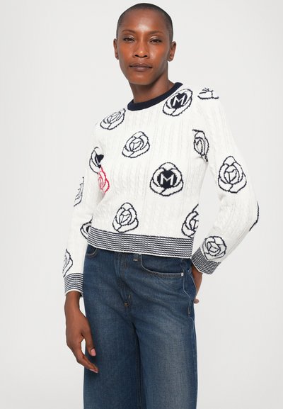 Frau, die einen weißen Strickpullover mit schwarzen und roten Blumenmustern und dunkelblauen Jeans trägt, steht mit einer Hand hinter ihrem Rücken.