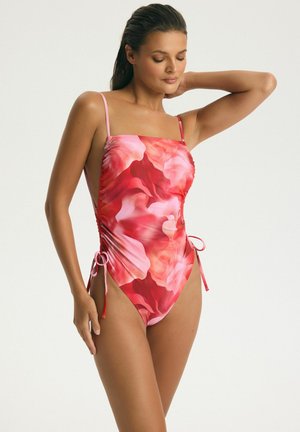 Žena v červeno-ružových kvetovaných jednodielnych plavkách s tenkými ramienkami a bočnými viazaniami, pózuje s jednou rukou zdvihnutou za hlavou.
