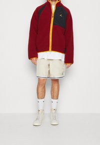Rote flauschige Jacke mit schwarzen Einsätzen, gelbem Reißverschluss, weißem T-Shirt, beigen Shorts mit schwarzen Akzenten, weißen Socken und hellen Schuhen.