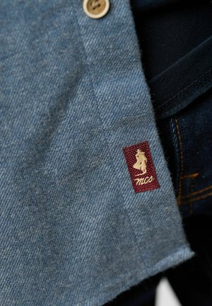 Camicia in denim di colore azzurro chiaro, con chiusura a bottoni e un'etichetta in tessuto bordeaux con un logo bianco ricamato sull'orlo.