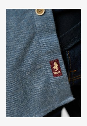 Jeanipaita vaaleansinisellä värillä, jossa on napitus, ja maroonivärinen kangastArréllä valkoinen logo brodeerattuna helmaan.