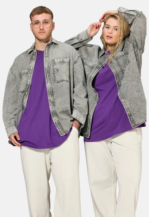 To personer iført grå denimjakker, lilla skjorter og hvide bukser står mod en ensfarvet lys baggrund.