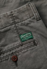 Tejido de mezclilla gris con un bolsillo de parche que presenta una etiqueta verde de "Superdry". Incluye un botón marrón con una marca grabada.