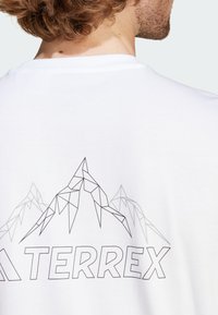 Bílá bavlněná tričko s geometrickým designem horského motivu v šedé barvě na zádech, s textem "TERREX" v tučném písmu pod ním.