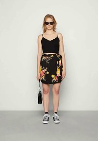 Mulher vestindo um top cropped preto com bordas em formato de concha, saia envolvente preta com flores, óculos de sol, tênis pretos, e segurando uma pequena bolsa preta, de frente.