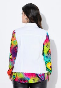 Chaqueta de denim blanca con vibrantes mangas de patrón de corazones multicolores, con bordes deshilachados y un cuello estructurado.