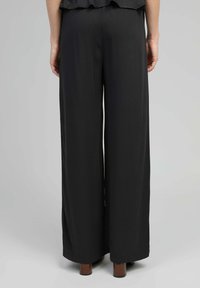 Vue de dos d'une personne portant un pantalon large noir flottant et des chaussures à talons carrés marron, se tenant contre un fond gris uni.