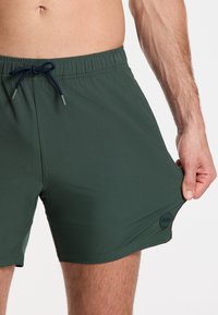 Zelené plávacie šortky vyrobené z ľahkej tkaniny, s elastickým pásom na šnúrku a malou nášivkou loga na boku.