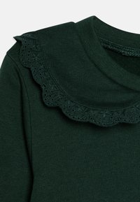 Indumento lungo verde scuro con maniche lunghe, caratterizzato da un collo rotondo e un bordo in pizzo scallop con dettagli a occhio lungo il colletto. Tessuto morbido e lavorato a maglia.