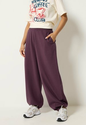Person trägt lockere violette Jogginghose mit elastischen Bündchen, weiße Sportschuhe und ein weißes T-Shirt mit roten und blauen Kaffee-Motiven.
