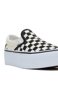 Baskets slip-on en toile à carreaux noirs et blancs avec une semelle en caoutchouc blanche, présentant des coutures contrastantes et une tige lisse et texturée.