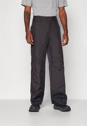 Hombre con pantalones impermeables negros de corte holgado, camiseta gris de manga corta y zapatos negros, de pie contra un fondo liso.
