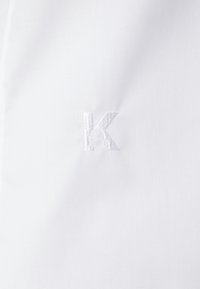 Wit stof met een subtiele, geborduurde letter 'K' in bijpassend garen. De textuur lijkt soepel en fijn te zijn.