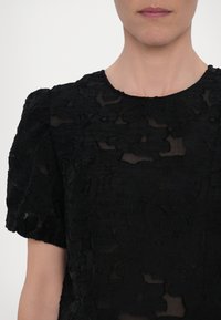 Abito nero con motivo floreale testurizzato, maniche a sbuffo corte e scollatura rotonda. Il tessuto appare trasparente con dettagli a strati.