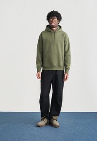 Πράσινη φούτερ μπλούζα με κουκούλα και μπροστινή τσέπη τύπου καγκουρό, συνδυασμένη με μαύρο παντελόνι. Το μοντέλο φοράει γκρι σουέτ παπούτσια με καφέ σόλες. Απλός σχεδιασμός, άνετη εφαρμογή.