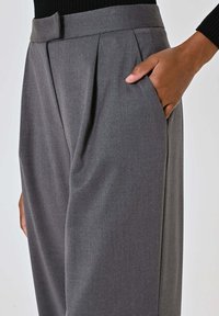 Pantaloni grigi a pieghe con una texture liscia. Dotati di una cintura in vita e tasche laterali, il tessuto appare leggero e formale.