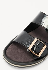 Sandale en cuir verni noir avec deux brides superposées, une boucle en or et une semelle intérieure en suede texturé. Équipée d'une semelle en caoutchouc avec des accents blancs.