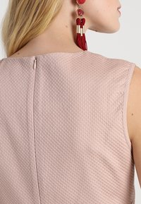 Donna bionda che indossa un abito senza maniche rosa pallido con texture, chiuso da una cerniera sulla schiena, e lunghe orecchini a frange rossi, con la testa inclinata di lato.