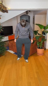 Fekete, laza szabású melegítőnadrág oldalzsebekkel, szürke cipzáras nyakú pulóverrel és ezüst tornacipővel párosítva. Beltéri környezet növényekkel.