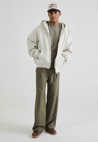 Felpa grigia chiara con zip, t-shirt verde oliva, pantaloni larghi verde oliva, scarpe marroni e un cappellino bianco con testo rosso. Outfit informale con texture morbide.