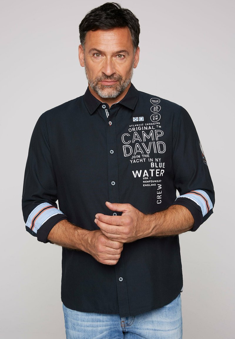 Camisa de botonadura en azul marino con bordados y parches en blanco, con acentos de rayas en las mangas dobladas y un corte ajustado.