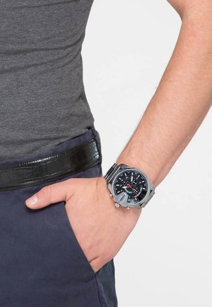 Silver stainless steel watch with a black dial, red accents, and textured band, worn on a hand resting in a navy pocket.