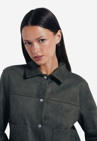 Oliwkowa zamszowa kurtka z ostrym kołnierzem, dwiema przednimi kieszeniami i srebrnymi napami. Gładka faktura i proste detale wzornicze.