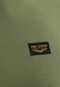 Tessuto di maglietta verde oliva con una superficie liscia e una toppa nera rettangolare che mostra "PME LEGEND AMERICAN CLASSIC" in caratteri dorati e ali.