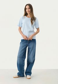 Camiseta de manga corta azul claro, de corte relajado, combinada con jeans de mezclilla azul de pierna ancha y zapatillas beige, sobre un fondo blanco liso.