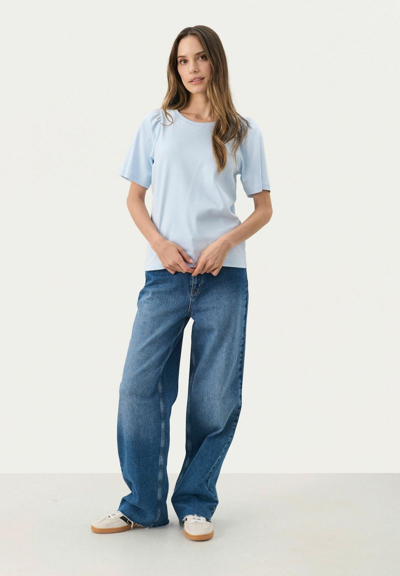 Camiseta de manga corta azul claro, de corte relajado, combinada con jeans de mezclilla azul de pierna ancha y zapatillas beige, sobre un fondo blanco liso.