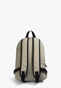Mochila de tela beige con correas acolchadas negras, un asa de transporte superior y un bolsillo lateral con cremallera. Textura suave con un sutil detalle de costura.