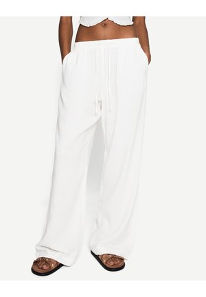 Personne portant un pantalon blanc ample avec cordon de serrage et poches ainsi que des sandales marron, debout devant un fond clair uni.