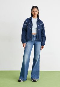 Casaco cortavento navy com costuras brancas, fecho de correr na frente e punhos elásticos, combinado com uma camisa curta azul clara e jeans largos desbotados.