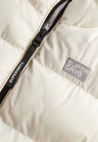 Giacca imbottita color crema con zip nera, caratterizzata da un grande logo ricamato grigio e texture trapuntata.