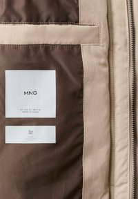 Beżowa tkanina o gładkiej fakturze, z białą metką oznaczoną napisem "MNG", wskazującą rozmiar i instrukcje dotyczące pielęgnacji oraz brązową podszewką.